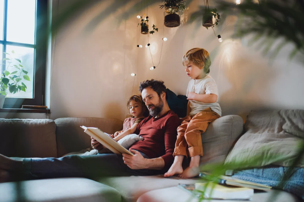 Father reading a book to two young children on a cozy couch, surrounded by plants and soft lighting