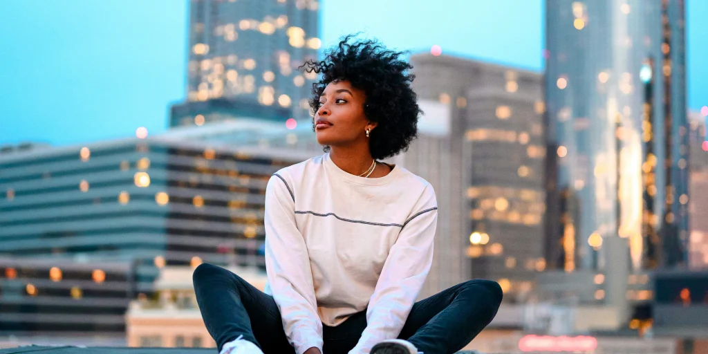 Woman with curly hair sitting on a rooftop, city skyline with lights in the background as the sun sets