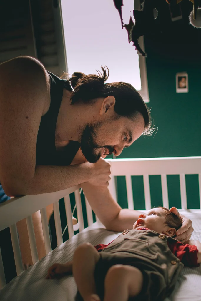 A man with long hair gently looks at a baby lying in a crib, creating a warm and tender moment